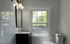 Bright bathroom with a large window, elegant vanity with gold faucet, and white marble tiles.
