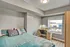 Cozy bedroom with a soft turquoise bedspread, a sleek wooden desk, and a large window offering a vibrant cityscape view.