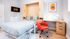 Bright modern bedroom with a wall-mounted TV, cozy bed with wooden frame, and stylish workspace featuring a vibrant red chair and sleek desk.