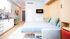 Modern studio apartment with a cozy bed, sleek kitchen, and vibrant green dining chairs, featuring contemporary artwork and a minimalistic design.