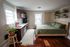 Cozy studio apartment featuring a stylish kitchenette with rich wood cabinets, a comfortable bed with green linens, and charming decor including plants and a woven wall hanging.