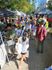 Vibrant outdoor market bustling with visitors exploring various vendor stalls under shady trees.