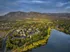 Breathtaking aerial view of a serene residential area surrounded by lush greenery, shimmering lake, and majestic mountains in the background, offering a perfect blend of nature and comfort.
