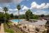 Serene apartment pool area under a vibrant sky, featuring palm trees, manicured gardens, and elegant poolside seating.