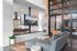 Modern living area with sleek grey sofa, stylish mounted TV, and contemporary kitchen in the background. Brick walls and large windows provide an airy and sophisticated atmosphere.
