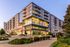 Modern multi-story apartment building with large windows and balconies, featuring ground floor retail space and vibrant landscaping.
