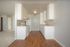 Bright and modern kitchen featuring sleek white cabinetry, spacious countertops, and stylish wood flooring, leading to a cozy carpeted living area.