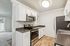 Modern kitchen with sleek stainless steel appliances, white cabinetry, and elegant wood flooring leading to a bright living space with a sliding glass door.