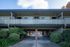 Charming two-story apartment building with spacious balcony and covered parking, surrounded by lush greenery on a sunny day.