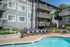 Charming apartment complex with a stylish poolside area, featuring comfortable lounge chairs and lush greenery under a bright blue sky.