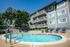 Charming apartment complex with a sparkling pool, surrounded by lush trees and cozy lounge chairs, offering a serene oasis for relaxation.