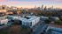 Breathtaking aerial view of modern apartment complex nestled in a vibrant urban neighborhood, surrounded by lush greenery and a stunning city skyline under a colorful sunset.