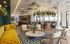Chic and inviting open-concept living space featuring a vibrant mustard sofa, elegant seating area, stylish bar with hanging greenery, and expansive windows offering scenic views.