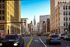 Dynamic city street view with bustling traffic, surrounded by towering architectural buildings, leading to a striking historic clock tower under a clear blue sky.