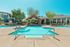 Inviting apartment pool area with crystal-clear water, modern lounge chairs, and a cozy pergola under a sunny blue sky, surrounded by lush greenery.