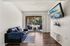 Bright living room with sleek hardwood floors, cozy black sofa, mounted flat-screen TV, and sliding doors leading to a sunny balcony with seating, offering a serene outdoor view.