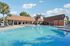Luxurious swimming pool area with crystal-clear water, surrounded by modern lounge chairs and a stylish clubhouse under a bright blue sky.