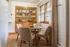 Charming dining area with elegant wooden shelves, cozy round table, and comfortable chairs, bathed in natural light from large windows.