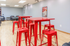 Vibrant communal area with striking red high tables and stools, perfect for social gatherings. Bright, spacious room with hardwood floors and modern decor.