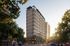Modern high-rise building with sleek architecture and a vibrant urban setting, surrounded by lush greenery and lively city streets.