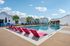 Inviting outdoor pool area with vibrant red lounge chairs and charming residential cottages in the background under a bright blue sky.
