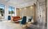 Contemporary lounge space with vibrant orange sofa, elegant blue chairs, and artistic partition wall, bathed in natural light from large windows.