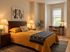 Inviting bedroom with warm lighting, featuring a cozy yellow quilted bed, elegant wooden furniture, and a chic desk nook by a bright window.