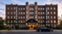 Elegant brick apartment building with lush greenery, featuring 'Baker House' and 'Now Leasing' banners, warmly lit entrance at dusk.