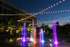 Vibrant and colorful water fountains lit up at night, with string lights overhead, set in an outdoor environment.