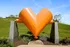 Vibrant orange heart-shaped sculpture standing against a lush green hill with a clear blue sky, creating an eye-catching outdoor installation near Yard 56 Lofts.