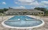 Inviting community pool with crystal-clear water, surrounded by a modern clubhouse and lush greenery under a bright blue sky.