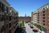 Vibrant urban courtyard framed by elegant brick apartment buildings under a clear blue sky, featuring a bustling parking area and charming shops.