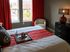 Cozy bedroom with modern decor, featuring large windows with red curtains, a stylish dresser, and a comfortable bed with decorative pillows.
