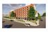 Modern urban apartment building with vibrant red brick facade, outdoor cafe seating, and lush green surroundings on a sunny day.