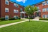 Charming brick apartment building with green manicured lawns, inviting pathways, and lush trees under a clear blue sky.