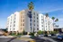 Contemporary six-story apartment building with sleek gray and beige exterior, surrounded by palm trees and city street in sunny weather.