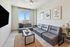 Bright and modern living room with sleek gray sofas, large window with city views, stylish decor, and fresh flowers on a side table.