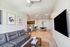 Modern apartment living room featuring sleek gray sofa, wall art, and open-plan kitchen with bar stools and dining area.