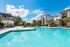 Luxurious apartment complex with a sparkling swimming pool, surrounded by modern buildings and lush greenery under a bright blue sky.