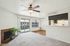 Charming living room with a cozy fireplace, elegant ceiling fan, and bright sliding glass doors leading to a balcony. Features plush carpeting, a stylish open kitchen, and inviting natural light.