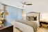 Bedroom with a king-sized bed, side table, dresser, beige carpeting, large window, and ceiling fan.