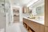 Bathroom with white marble tile on the floors and shower walls, minimalist wood cabinets, a dual vanity with a white quartz countertop, and an attached walk-in closet with beige carpeting.