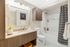 Bathroom with white marble tile on the floors and shower walls, minimalist wood cabinets, an ecru-colored quartz countertop, and a full-sized soaking tub with a shower curtain.