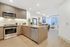 Open-concept kitchen and living space with ash-gray wood cabinets and light wood-style floors, tan quartz countertops, a peninsula with an undermount sink, stainless steel appliances, and a spacious living room with a raised ceiling.
