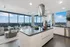 Open-concept living, dining and kitchen space with dark wood-style floors, a large island with an electric range and stainless steel overhead hood, white handleless cabinets, and a panoramic city view through two full-width floor-to-ceiling windows.