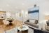 Open-concept living and dining area with a couch, side table, armchair, and coffee table in the foreground and a 4-person dining table beyond. A large blue and white rug is on the floor, which matches the colors in the abstract landscape on the wall.