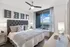 Bedroom with king-sized bed, dresser, white walls, dark wood-style floors, ceiling fan, and large window overlooking a balcony and a view of the park.