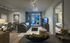 Living room with a floor-to-ceiling window opening to a balcony, light brown wooden floors, modern silver track lights, dark gray and beige couch and chairs, TV, and a doorway leading to a bedroom.