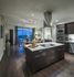 Open kitchen and living area with floor-to-ceiling windows, dining table with orange chairs, grey countertops and brown cabinets. There is a large kitchen island in with an electric stove, vent hood, and double wall ovens.