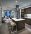 A kitchen with dark wood-style cabinets, stainless steel appliances, and a white quartz peninsula with 3 bar stools. There is an open living room and dining area with floor-to-ceiling windows.